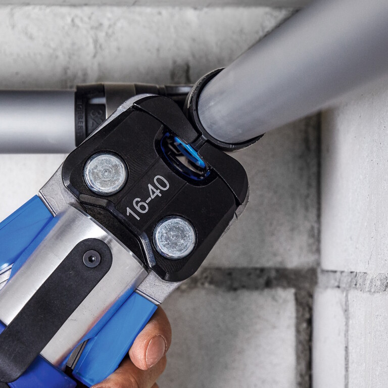 Eine Hand hält ein Presswerkzeug, das zwei an einer weiss gestrichenen Ziegelwand verlaufende Rohre verbindet.