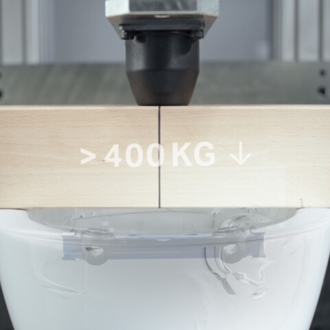 Eine Maschine drückt zu Testzwecken einen Holzbalken auf ein weisses, wandmontiertes WC. Auf dem Holzbalken ist «> 400 kg» aufgedruckt.