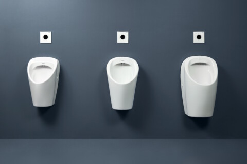 Drei weisse Urinale sind in einer Reihe an einer dunkelgrauen Wand montiert.