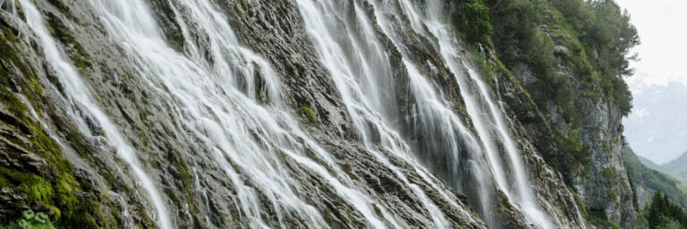 Mehrere Wasserfälle rauschen eine steile, felsige Felswand hinab, umgeben von grüner Vegetation.