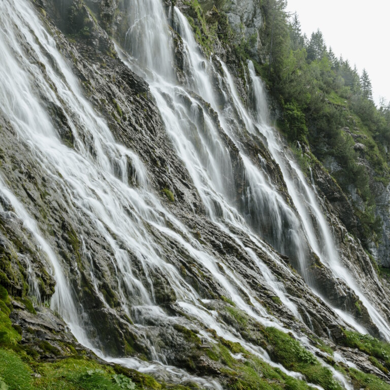 Mehrere Wasserfälle rauschen eine steile, felsige Felswand hinab, umgeben von grüner Vegetation.